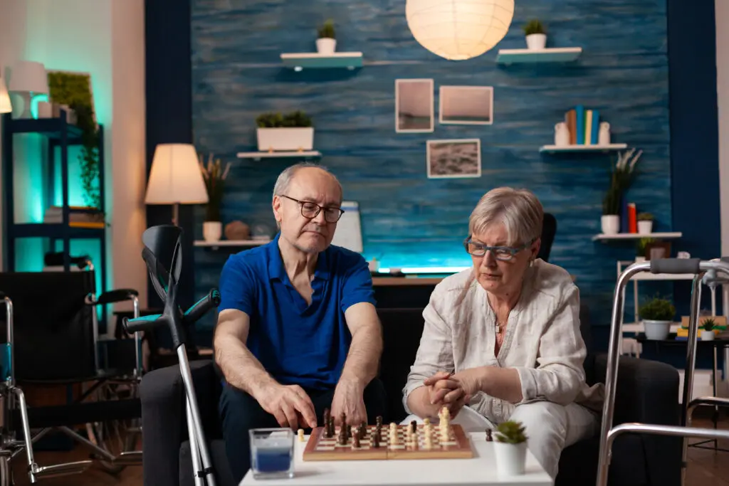 &Auml;lteres Paar spielt gemeinsam Schach im Wohnzimmer, Gehhilfe und Rollator stehen griffbereit daneben.